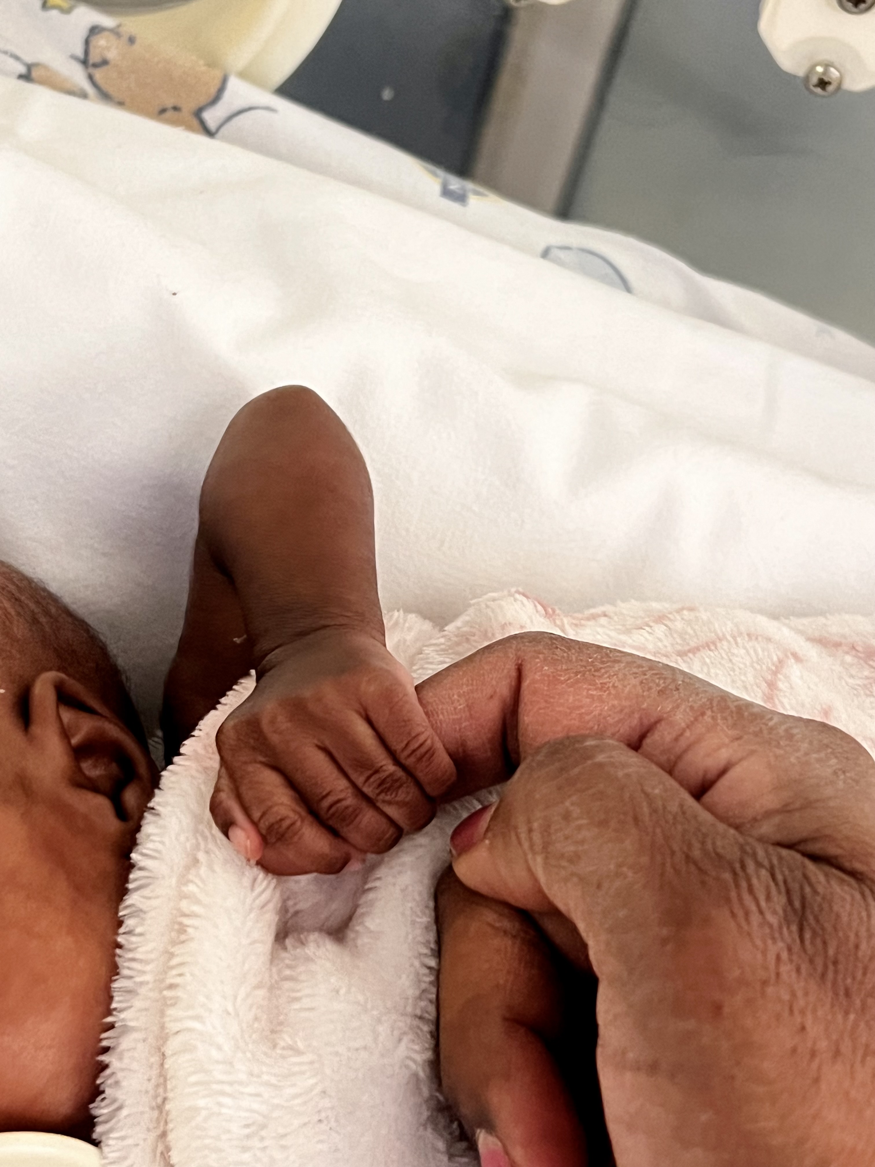 A newborn baby's tiny hand gently grasping Dr. Vinola Naidoo’s finger, wrapped in a soft blanket in a hospital setting.