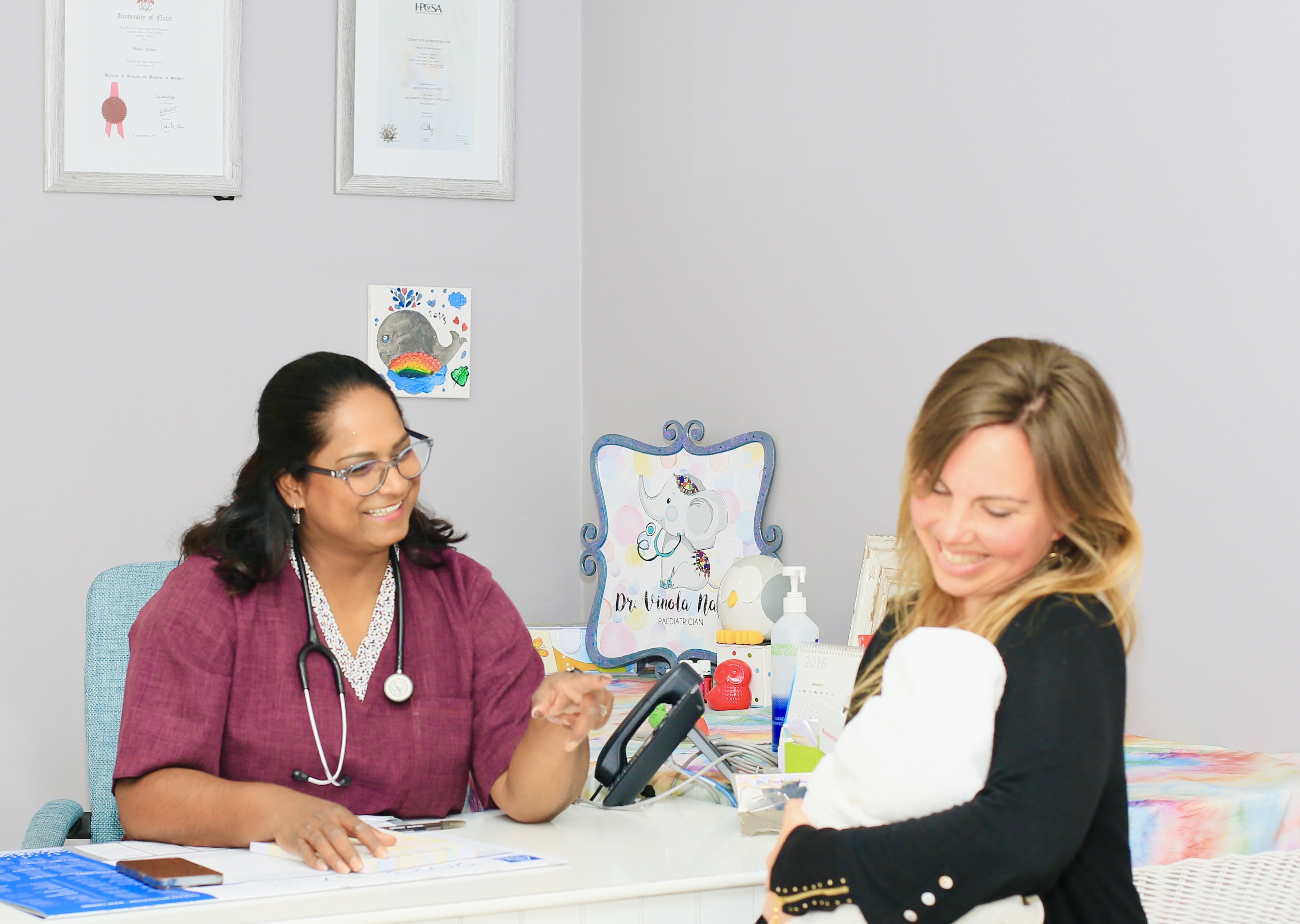 Dr. Vinola Naidoo warmly speaking to a smiling mother holding her baby during a consultation at her desk.