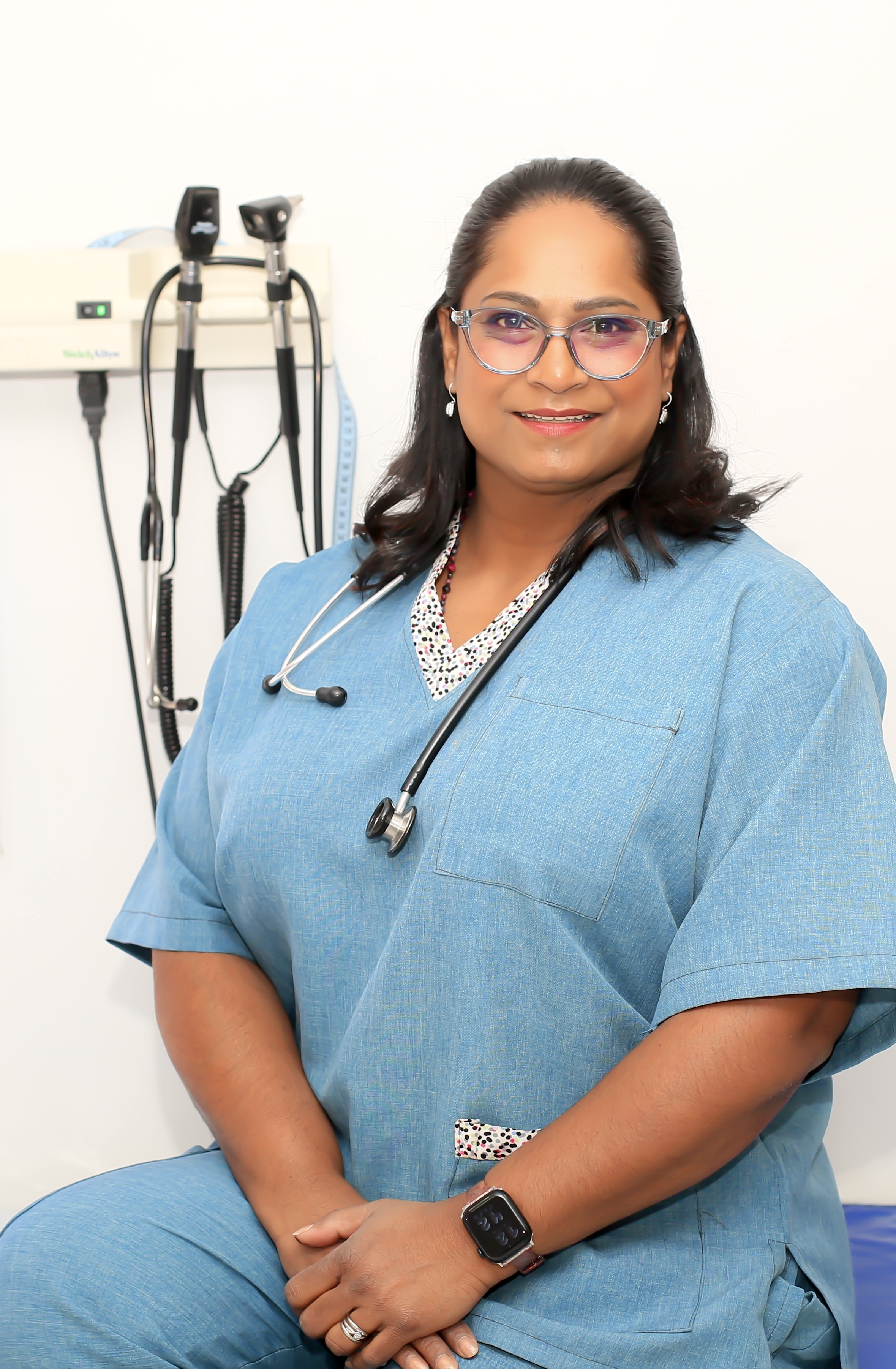 Dr. Vinola Naidoo smiling confidently in blue scrubs with a stethoscope, standing in a bright clinical room with medical tools in the background.