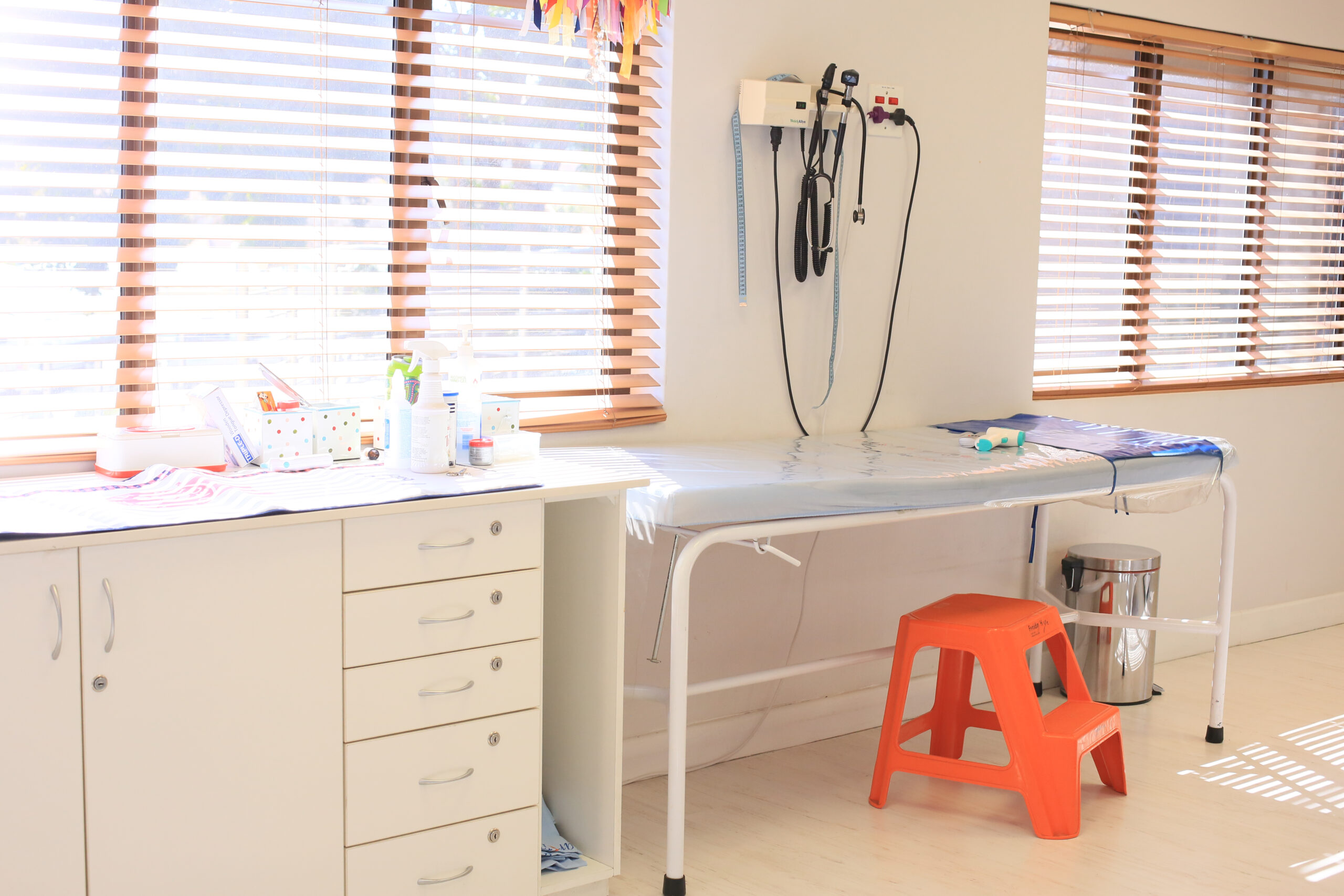 Examination room with a clean medical bed, pediatric tools on the counter, and a bright orange step stool under natural sunlight.
