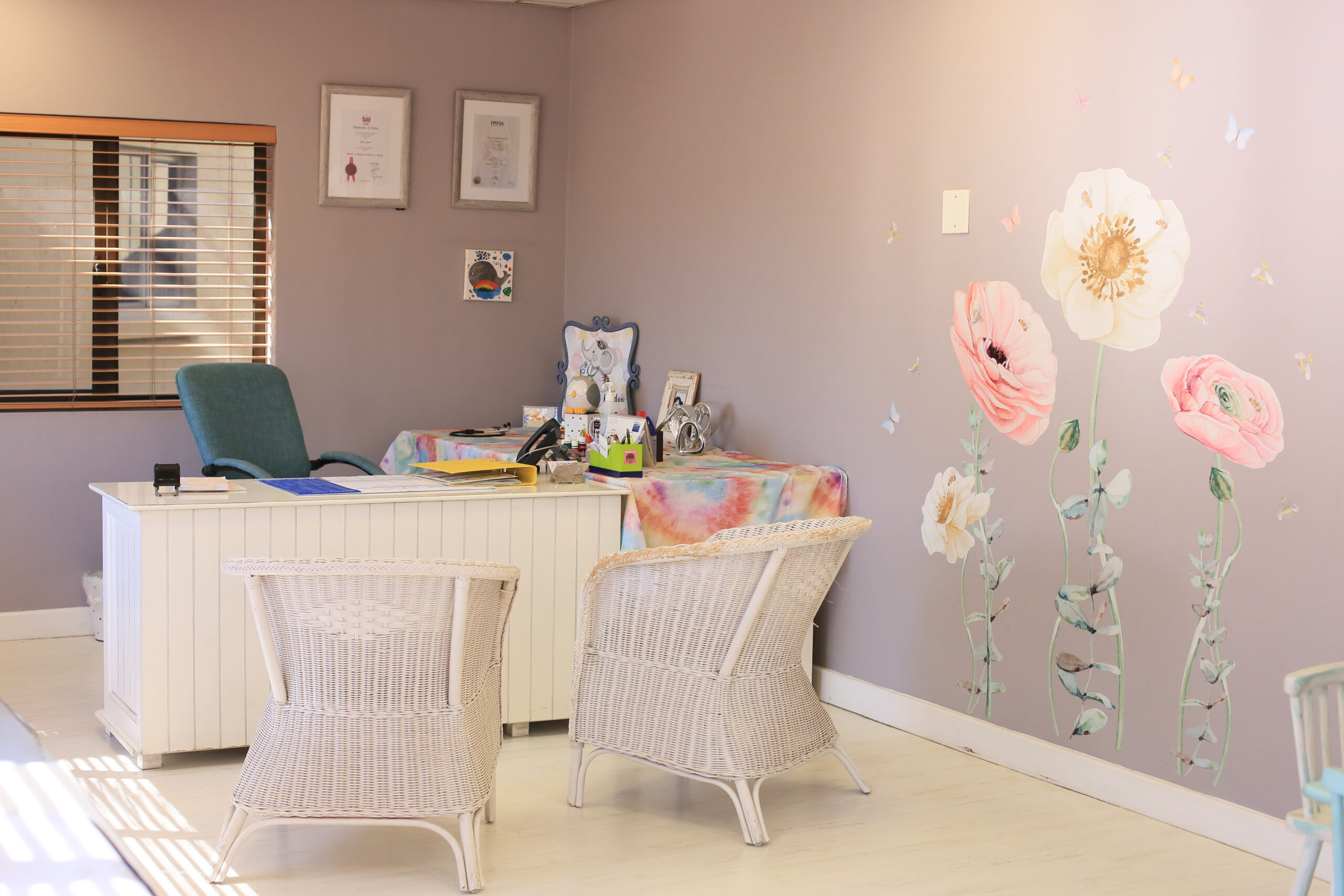 Cozy pediatric office with white wicker chairs, a desk covered in a colorful cloth, certificates on the wall, and hand-painted flower murals.