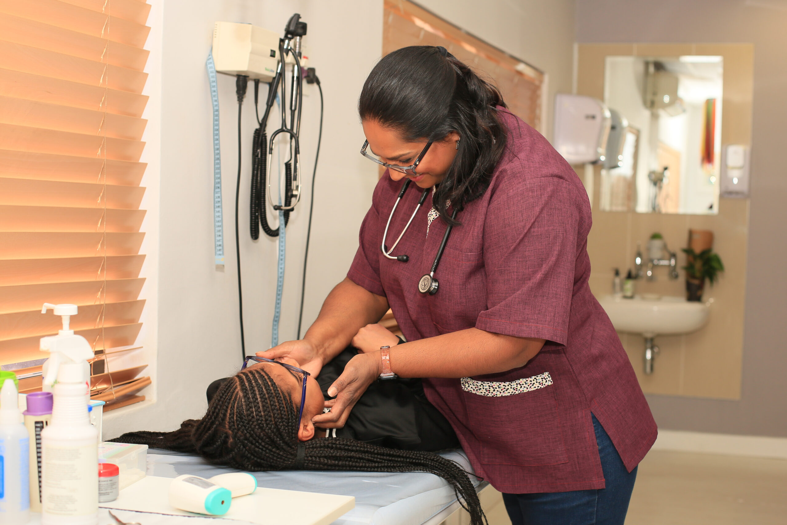 Dr. Vinola Naidoo gently examining a child, wearing a stethoscope, surrounded by medical equipment.