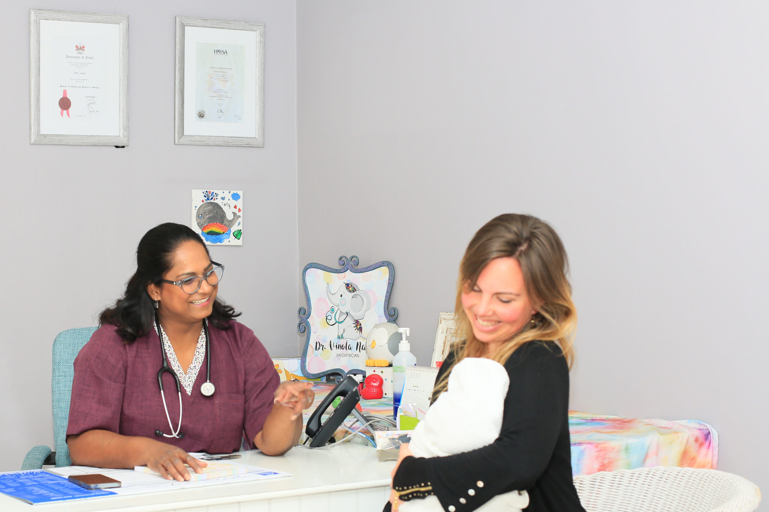 Dr. Vinola Naidoo warmly speaking to a smiling mother holding her baby during a consultation at her desk.