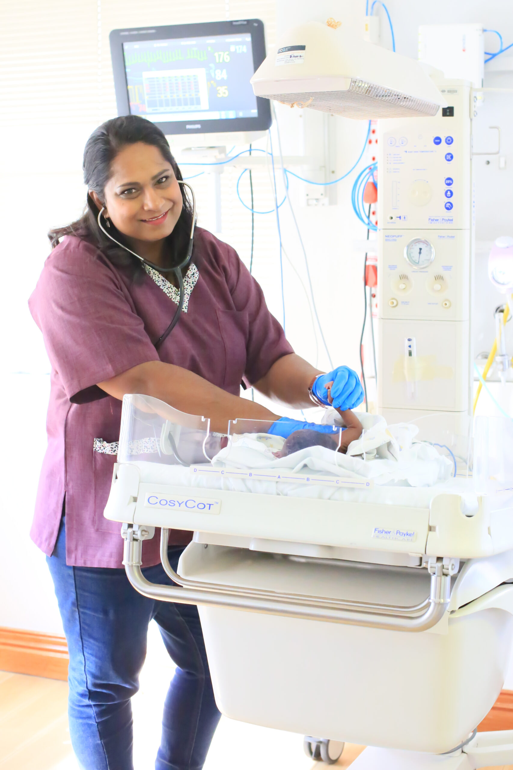 Dr. Vinola Naidoo caring for a newborn inside a neonatal CosyCot unit, monitoring vital signs during a hospital check-up.