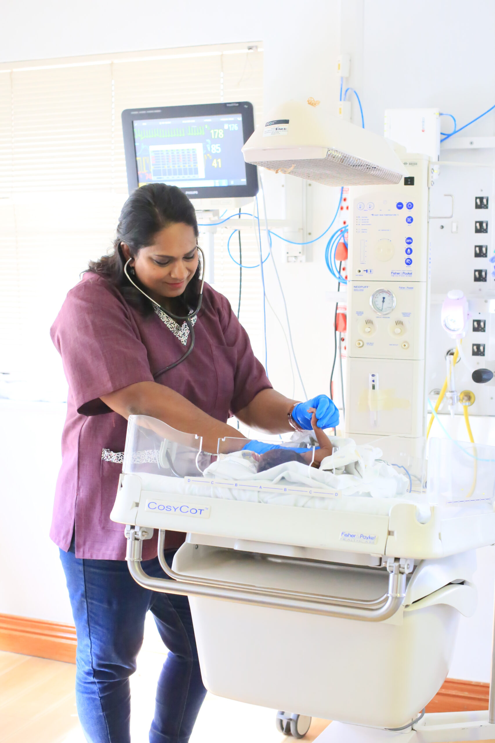 Dr. Vinola Naidoo gently examining a newborn’s hand in a neonatal unit, wearing gloves and a stethoscope, surrounded by medical equipment.