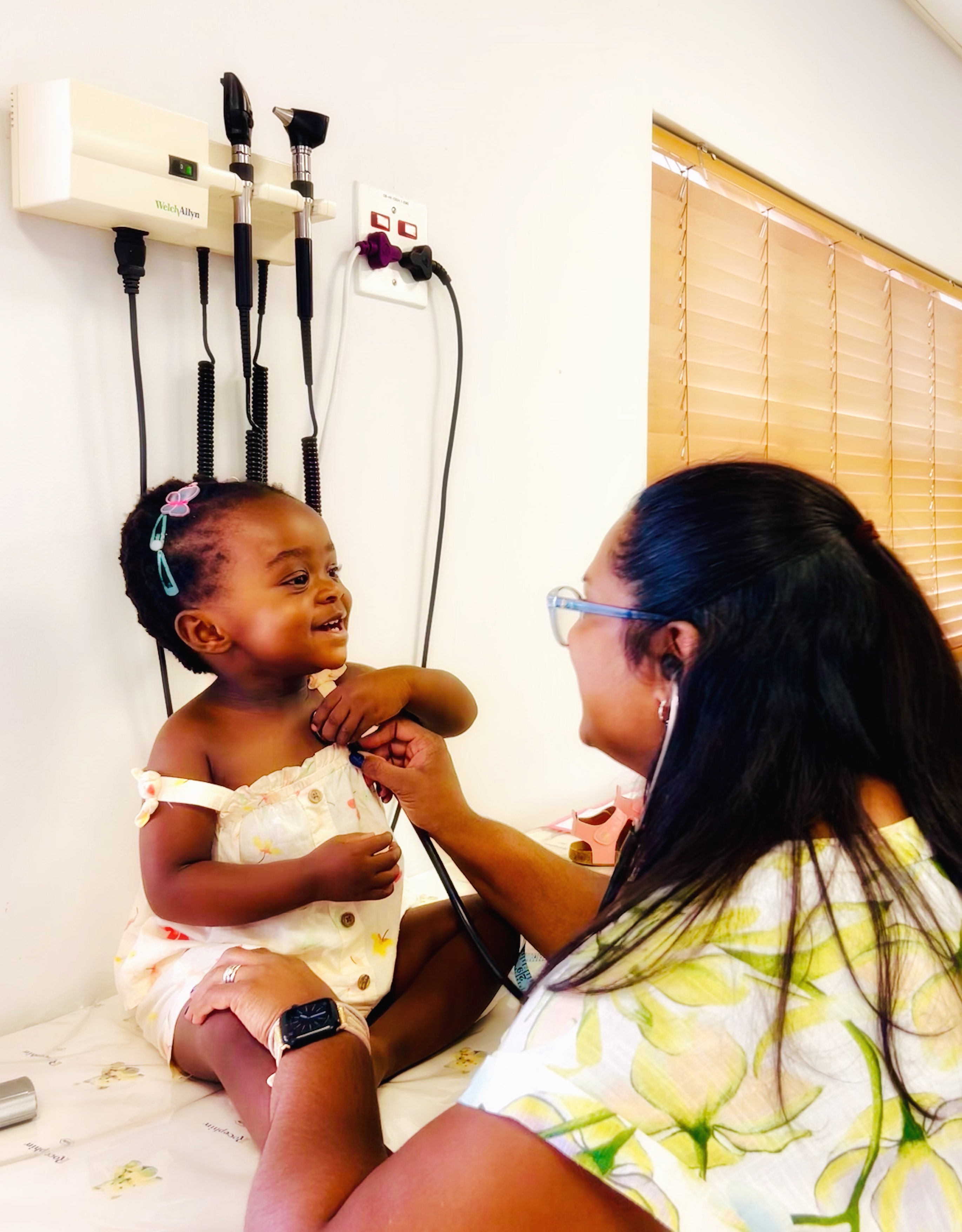 Dr. Vinola Naidoo listening to a joyful toddler’s heartbeat with a stethoscope during a cheerful check-up on an examination bed.